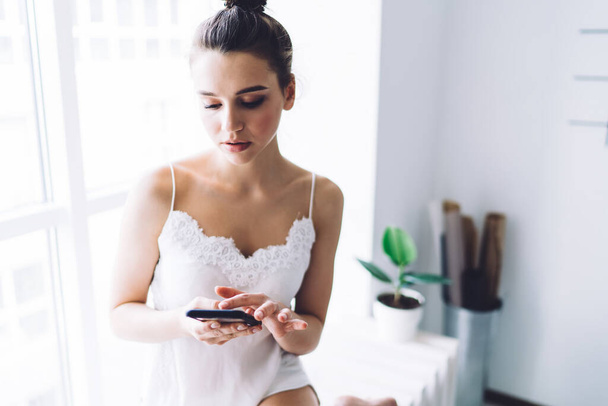 Young calm attractive woman in pyjamas browsing smartphone while sitting at window sill in urban apartment with white walls in morning - Fotoğraf, Görsel