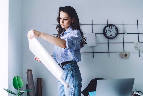 Side view of thoughtful young lovely brunette female in blue holding and looking at unrolled white craft paper while standing at window in room - Foto, Imagem