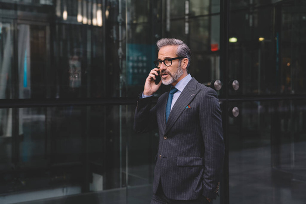 Middle aged businessman with beard in stylish dark business suit talking on phone near office building putting hand in pocket in New York - Photo, Image