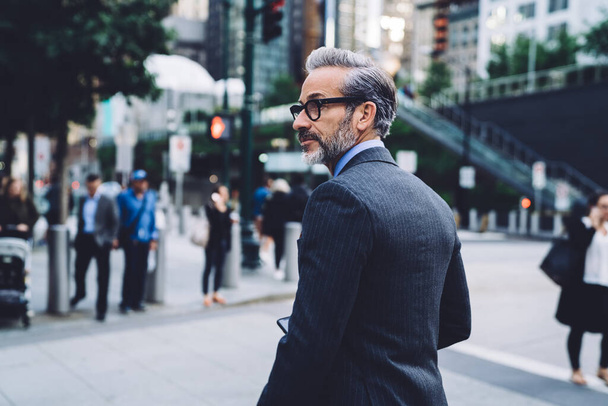 Half turned fashionable mature businessman in glasses and black suit looking away on busy street in New York on blurred background - Fotografie, Obrázek
