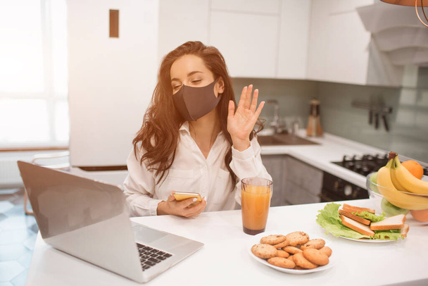 Vanwege de coronavirus pandemie, een vrouw in isolatie thuis. Ze werkt thuis, draagt een masker en heeft een videoconferentie op haar laptop. - Foto, afbeelding
