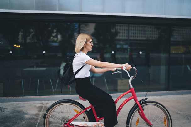 Side view of attractive female student with blonde hair in casual clothing with backpack riding bike with red frame smiling and looking frontwards - Foto, Bild