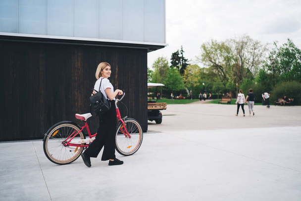 Side view of pleased adult cyclist blond female in casual outfit and with black backpack walking with bicycle against street building looking away  - Fotografie, Obrázek