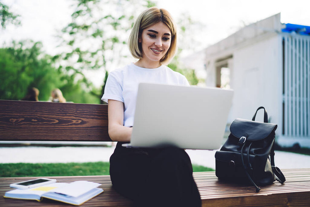 Cheerful and positive female freelancer with laptop sitting on wooden bench in park and smiling while texting in social media - Фото, изображение
