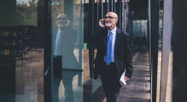 Confident old businessman in suit and eyeglasses and with white beard holding newspaper having phone call while walking in passage on street looking away - Photo, Image