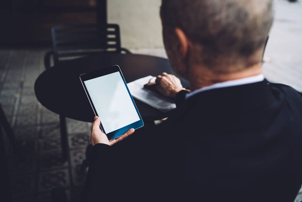 Homme d'affaires respectable âgé assis avec tablette noire devant la table ronde dans un café accueillant et naviguant sur les médias sociaux pendant la pause
 - Photo, image