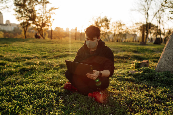 Przystojny brodaty mężczyzna siedzi na trawie na słońcu z laptopem. Maska na twarz. Koronawirus - Zdjęcie, obraz
