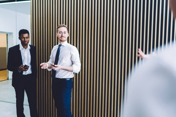 Multiracionális fókuszált sikeres menedzserek okostelefonnal és kollégákkal beszélgetnek, miközben a céges irodafolyosó szünetében a csíkos falnál állnak  - Fotó, kép