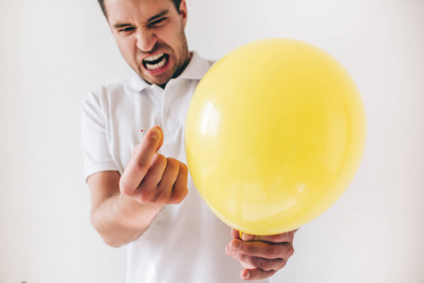 Young man isolated over white background. Guy emotional expressing before piecercing a yellow balloon with needle. Cut view. - Photo, Image