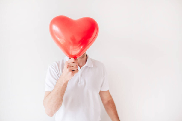 Mladík izolovaný na bílém pozadí. Ten chlap drží červený balón ve tvaru srdce před obličejem. Zakryj nebo skryj. Láska. - Fotografie, Obrázek