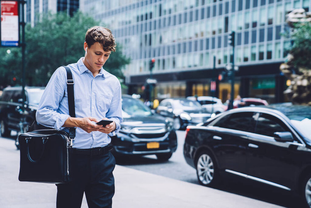 Male handsome employee in formal wear and wireless headphones with big leather bag messaging on phone walking down street of New York city - Фото, зображення