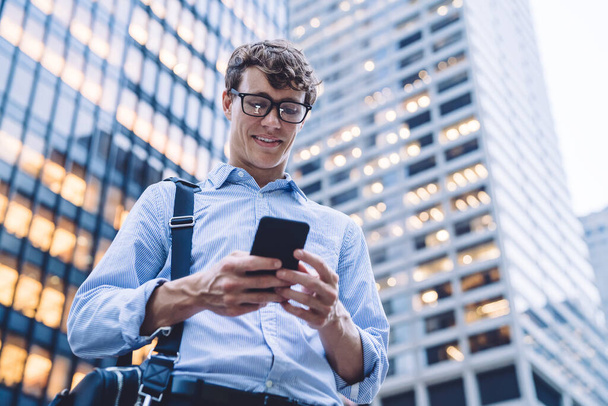 Desde abajo joven guapo hombre feliz en gafas con ropa de oficina usando un teléfono inteligente moderno en la calle en frente del edificio de oficinas en Manhattan, Nueva York
 - Foto, Imagen