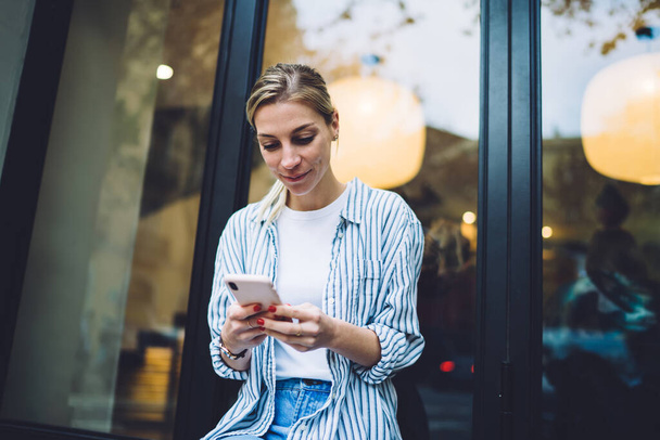 Trendy blond haired young woman in white T-shirt under striped shirt and blue jeans browsing on smartphone on street against glass window - Photo, Image