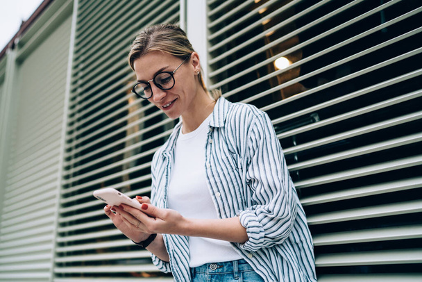 Сверху молодая радостная леди в повседневной одежде просматривает социальные сети на смартфоне и улыбается, опираясь на окно вслепую
 - Фото, изображение