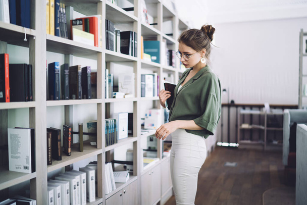 Young attractive woman with digital tablet in hands standing in college library in front of bookshelves reading titles and searching book on daytime - Φωτογραφία, εικόνα