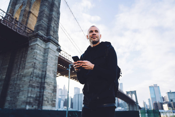 Glad smiling short haired young man in black sportswear with smartphone and backpack standing near massive bridge in daylight on urban background and looking away - Foto, afbeelding