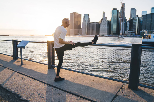 Adult athletic man in sportswear warming up while listening to music in earphones on embankment of river on urban background with skyscrapers - Foto, immagini