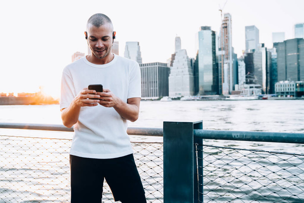 Young ethnic man in white shirt with earphones using smartphone and standing near metallic fence with chain link and New York river against background of city - Foto, Imagem