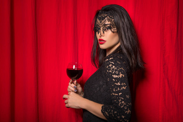 Young beautiful woman holding glass of wine over red curtains - 写真・画像