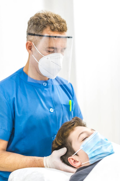 Physiotherapist with mask and screen giving a cranial massage to a child with a mask. Physiotherapy safety measures in the Covid-19 pandemic. Osteopathy, therapeutic chiromassage - Foto, imagen
