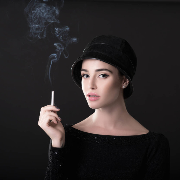 Young fashion woman smoking cigarette in hat and black drees over dark background. Vintage female portrait, styling. Image toned. - 写真・画像