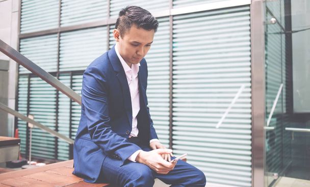 Side view of young Asian businessman dressed in blue formal suit sitting on bench with pensive face while chatting on social network - Foto, Imagem