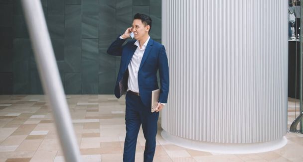 Delighted successful handsome Asian young man in suit holding laptop while standing and talking over phone beside urban office building in daylight - Фото, изображение