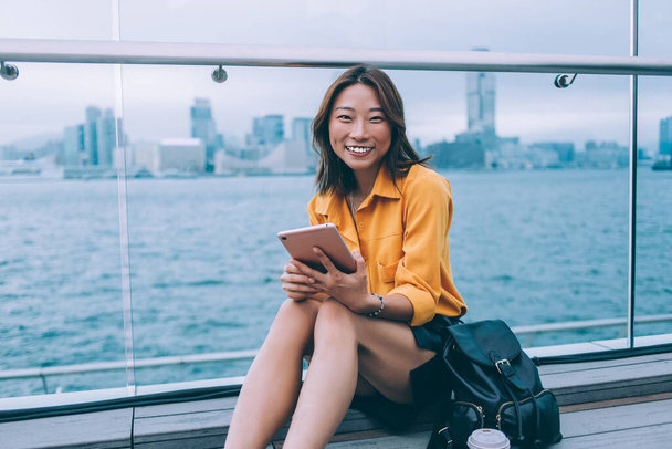 Happy young Asian woman in casual clothes with leather backpack sitting on Hong Kong wharf and holding tablet smiling at camera  - Фото, зображення