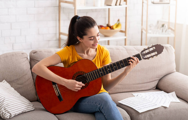 Μείνε σπίτι για διασκέδαση. Millennial κορίτσι μαθαίνουν να παίζουν ακουστική κιθάρα στο διαμέρισμα - Φωτογραφία, εικόνα