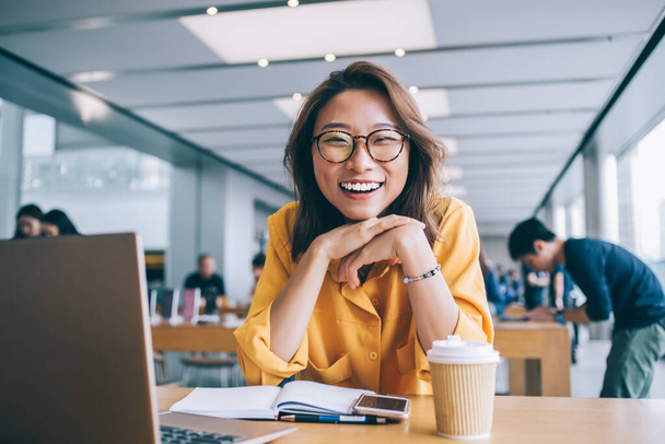 Beautiful happy Asian woman with glasses smiling widely looking at camera sitting at table in workspace in Hong Kong city with people in background - Photo, image