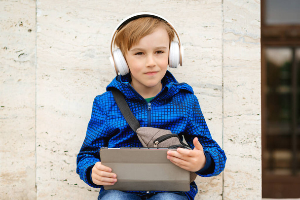 屋外のタブレットで音楽を聴いているかわいい少年。ワイヤレスヘッドフォンを使用して子供。ストリートを歩いて音楽を楽しむスタイリッシュな少年。人々、技術、ライフスタイル。外でいい子だ。ファッション. - 写真・画像