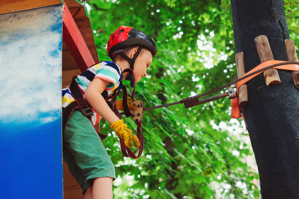 小さな男の子の木の間で高いケーブルルートを渡す。アドベンチャーパークの高いロープコースで子供クライミング。森のアドベンチャーパークの子供たち。子供は安全ヘルメット極端なスポーツ。子供のためのサマーキャンプ. - 写真・画像