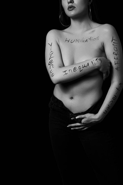 part of a young woman's body with the words affront,cruelty,inequality on a black background.photo.the inscription is not a tattoo, written by the photographer's hand. - Foto, imagen