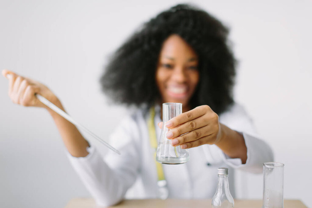 Young beautiful African American girl doctor in a white coat with a stethoscope. sitting at a table with reagent flasks on white background. Researcher researching in the laboratory - Zdjęcie, obraz