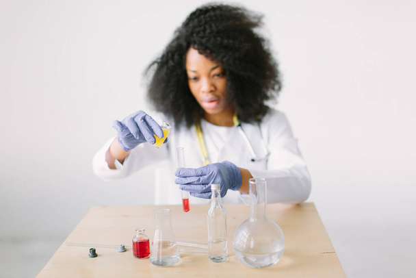 Young beautiful African American girl doctor in a white coat with a stethoscope. sitting at a table with reagent flasks on white background. Woman researcher doing a blood test in a laboratory - Fotó, kép