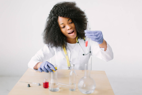 laboratory assistant analyzing a blood sample . Young beautiful African American girl doctor in a white coat with a stethoscope. sitting at a table with reagent flasks on white background. - Photo, image