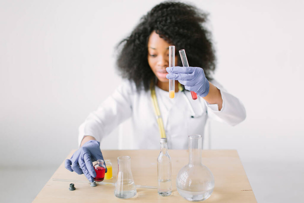 Young beautiful African American girl doctor in a white coat with a stethoscope. sitting at a table with reagent flasks on white background. Medical equipment. Blood test - Fotó, kép