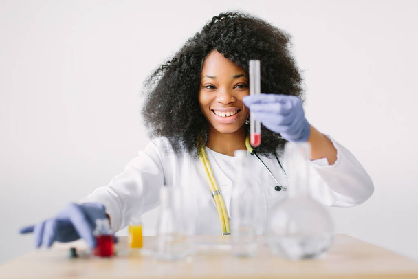 Young beautiful African American girl doctor in a white coat with a stethoscope. sitting at a table with reagent flasks on white background. Female laboratory assistant with blood test for AIDS - Φωτογραφία, εικόνα