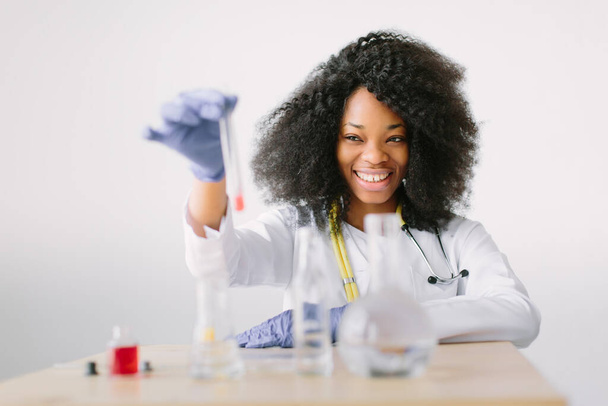 Young beautiful African American girl doctor in a white coat with a stethoscope. sitting at a table with reagent flasks on white background. Investigator checking test tubes. - Фото, зображення