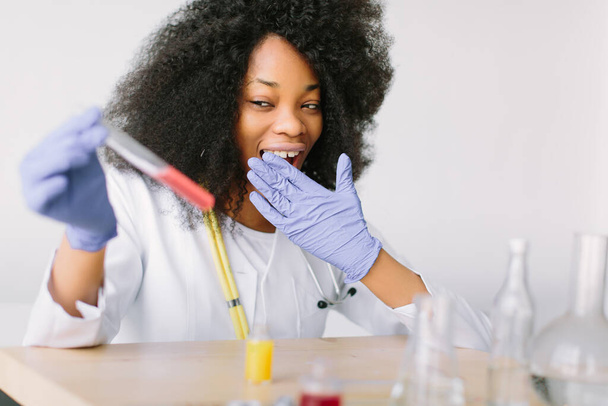 Young beautiful African American girl doctor in a white coat with a stethoscope. sitting at a table with reagent flasks on white background. Female lab assistant studying blood sample for analysis - Photo, image