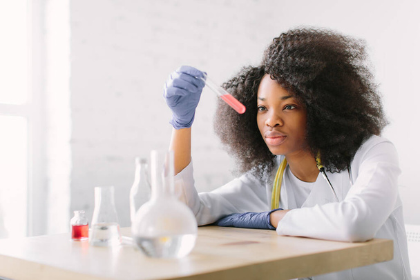 Jovem e bela menina afro-americana médica em um casaco branco com um estetoscópio. sentado a uma mesa com frascos de reagente sobre fundo branco. Assistente de laboratório estudando amostra de sangue para análise - Foto, Imagem