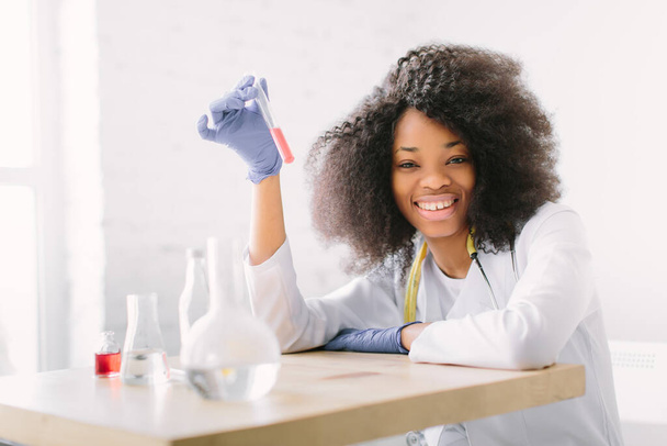 Young beautiful African American girl doctor in a white coat with a stethoscope. sitting at a table with reagent flasks on white background. Investigator checking test tubes. - Foto, Imagem