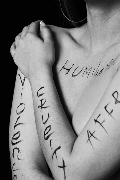 część ciała młodej kobiety ze słowami zniewaga, okrucieństwo, nierówność na czarnym tle.photo.the napis nie jest tatuażem, napisanym ręką fotografa. - Zdjęcie, obraz