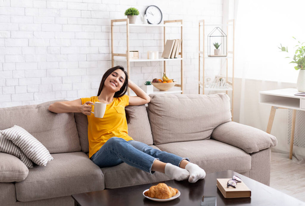 Nyugodt Y-generációs lány ül a kanapén egy csésze kávéval otthon, élvezi a békés reggelt - Fotó, kép