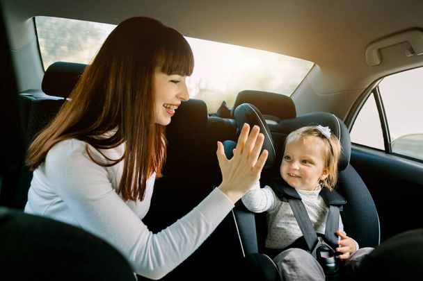 Pretty young woman mother takes care about her little daughter in a car safety seat, sitting together, giving high five each other and having fun. Safety road, happy family trip, baby safety - Φωτογραφία, εικόνα