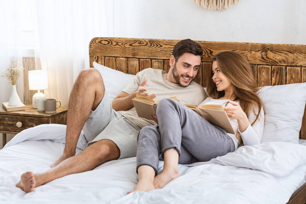 Couple reads books in bedroom. Guy and girl in home clothes on bed. - Φωτογραφία, εικόνα