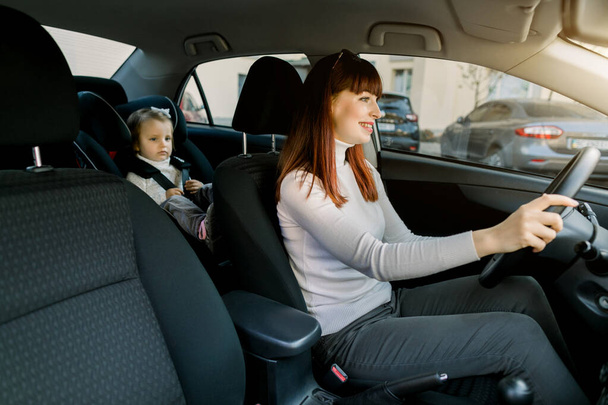 Pretty Caucasian woman, young mother driving a car, having her little baby girl in a child seat behind. Safety driving, children car seat concept - Photo, Image