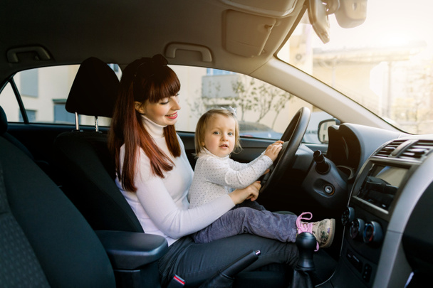 Молодая мать со своей маленькой девочкой, сидящей в машине на переднем сиденье возле руля автомобиля. Женщина и девочка, притворяющиеся, что они за рулем. Мама показывает своему ребенку машину внутри
 - Фото, изображение