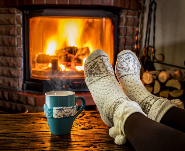 Pieds féminins en chaussettes de laine douce et feu brûlant dans la cheminée en arrière-plan. Se réchauffer et se détendre près de la cheminée avec une tasse de boisson chaude est la meilleure façon de se détendre et de se détendre après une période préoccupante ou occupée. Vacances d'hiver ou idées de Noël. - Photo, image