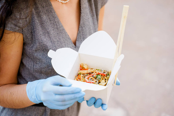 Closeup tasty food wok in box delivery. Girl holds udon noodles with tempuru, shrimps, soy sauce in hands in medical gloves. Japanese spicy food in disposable eco boxes. Wok box udon advertise - Photo, Image